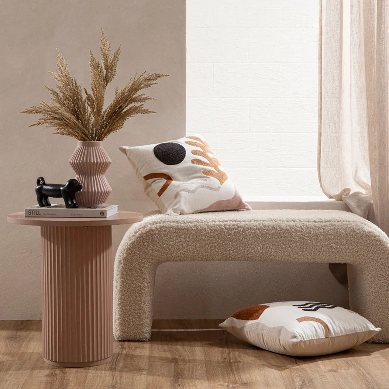 Decorative scene with a small table, vase with dried plants, and pillows on a bench.
