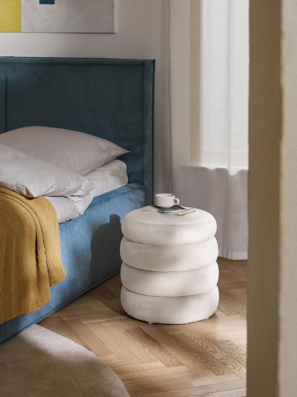 White cylindrical stool next to a bed with a blue headboard and yellow blanket.