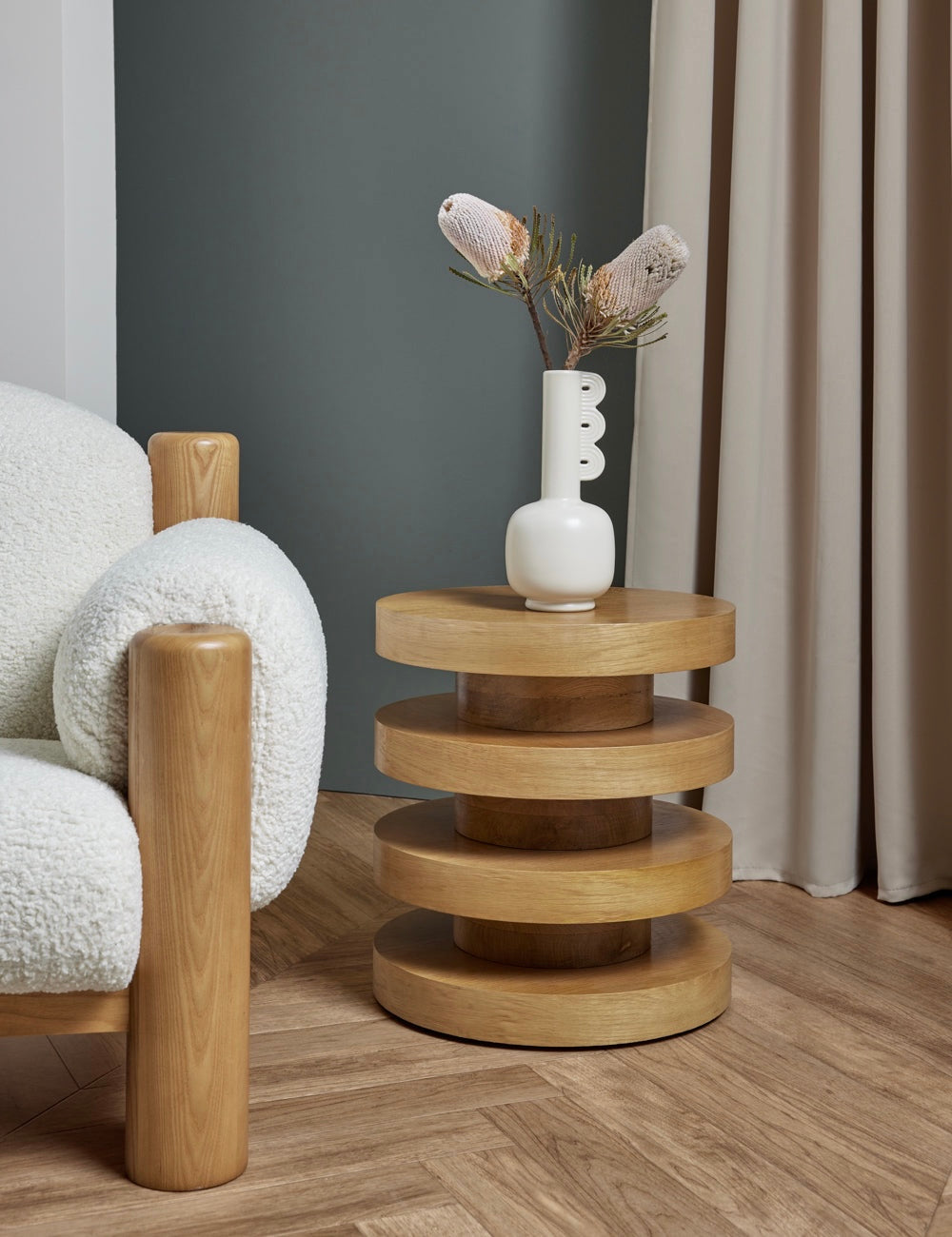 Wooden side table with a white vase and dried flowers next to a textured armchair.