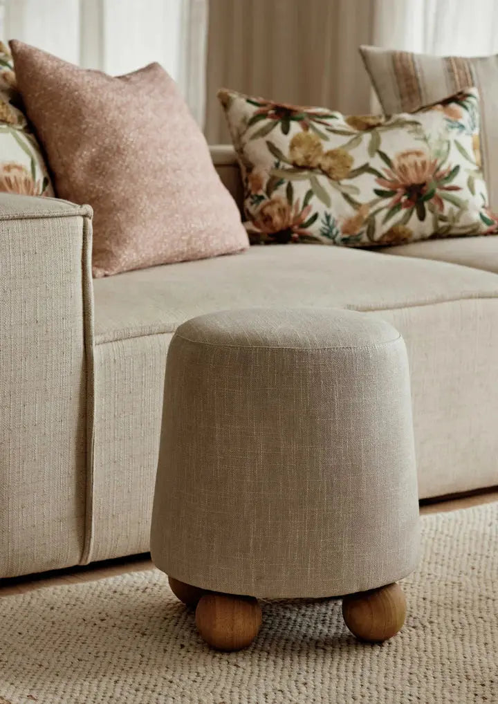 Beige ottoman with wooden legs in front of a beige sofa with patterned cushions.