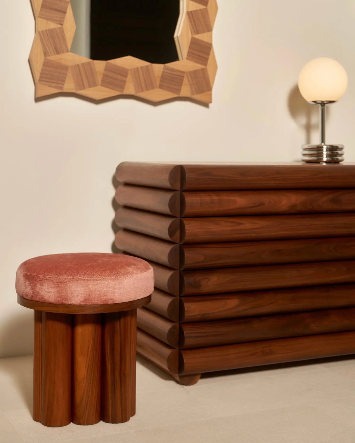 Wooden dresser, stool with pink cushion, and mirror with geometric frame in a room setting.
