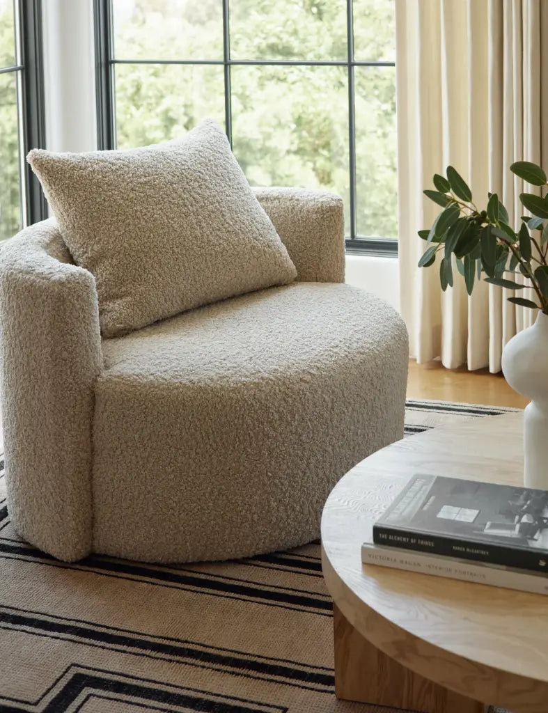 Beige armchair with a pillow in a living room setting with a coffee table and books.
