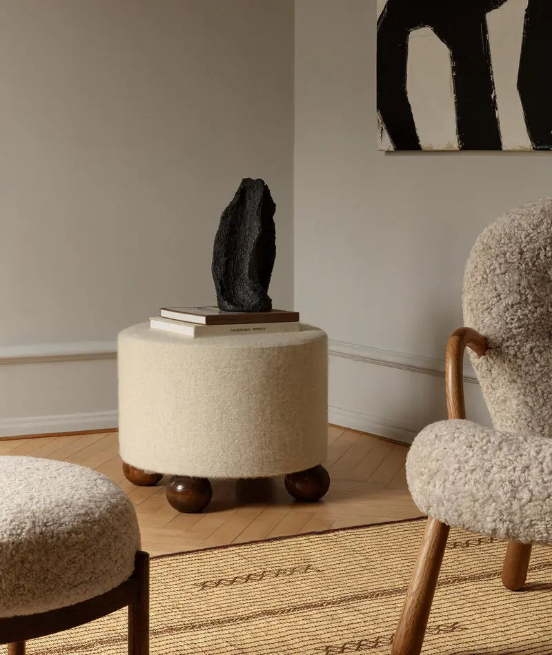 Round beige ottoman with a black sculpture on top, next to a textured chair in a room setting.