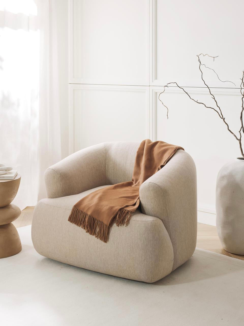 Beige armchair with a brown blanket in a minimalistic room with white walls and light wood flooring.