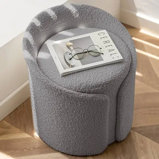 Gray plush ottoman with a magazine and glasses on top, placed on a wooden floor.